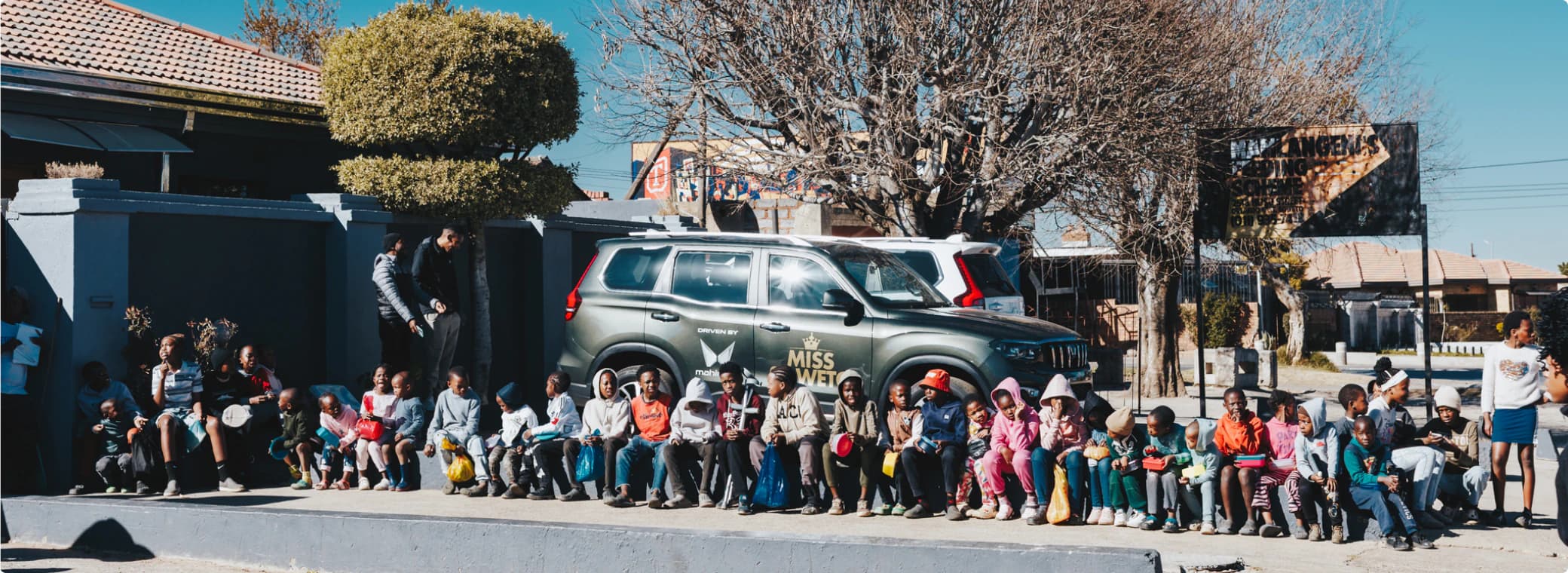 Children seated in a line beside a Mahindra vehicle at a Mandela Day community event