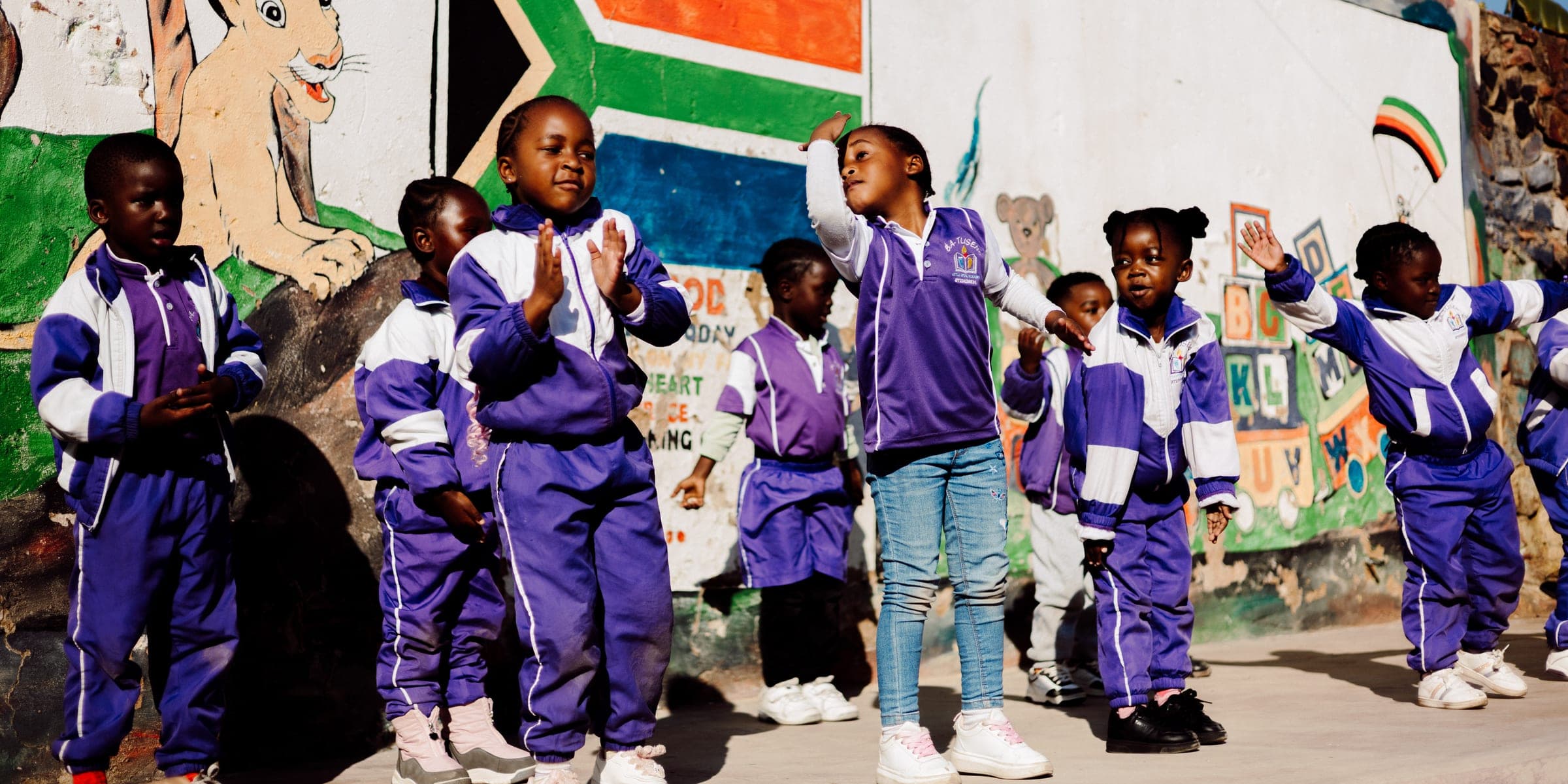Children in purple Ba-Tliseng tracksuits lined up in front of a mural wall